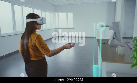 Kreative asiatische Designerin, die ein Augmented-Reality-Headset trägt, nutzt Virtual Interior Design Software und Controller, um einen Büroraum mit 3D-Möbeln und -Geräten zu bauen. Futuristische VFX-Animation. Stockfoto