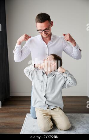 Junger Vater in Brille, der mit seinem neugierigen Sohn spielt. Lustiger Junge, der seinem Vater Muskeln zeigt. Familie genießt Aktivitäten zu Hause. Stockfoto