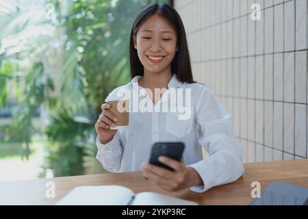 Eine fröhliche junge Frau sitzt an einem Holztisch, schlürft Kaffee und benutzt ihr Smartphone. Die Umgebung ist hell und mit Grün gefüllt, die einen Meter reflektiert Stockfoto