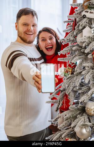 Mann mit Frau, die Telefon hält, mit leerem Bildschirm, der neben dem weihnachtsbaum steht Stockfoto