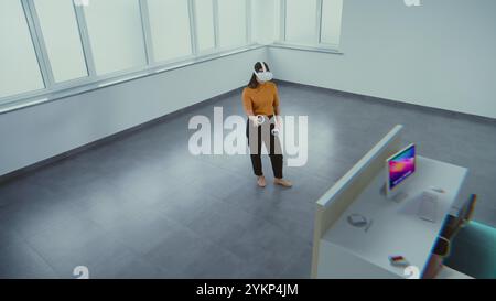Kreative asiatische Designerin, die ein Augmented-Reality-Headset trägt, nutzt Virtual Interior Design Software und Controller, um einen Büroraum mit 3D-Möbeln und -Geräten zu bauen. Futuristische VFX-Animation. Stockfoto