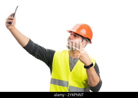 Erwachsener Baumeister Mann lächelt als Selfie mit Smartphone-Kamera, trägt orangefarbenen Schutzhelm und fluoreszierende Weste, die Frieden oder Siegeszeichen mit Flossen macht Stockfoto