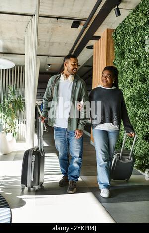 Ein fröhliches junges afroamerikanisches Paar schlendert durch eine lebhafte Hotellobby, mit Gepäck in der Hand. Stockfoto