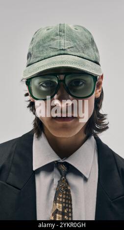 Nahaufnahme des Porträts eines stilvollen jungen Mannes mit grüner Mütze und übergroßer grüner Sonnenbrille, schwarzem Blazer, weißem Hemd und gemusterter Krawatte Stockfoto