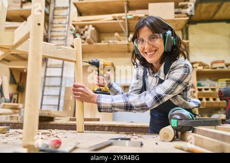 Eine lächelnde junge Frau arbeitet in einem Holzhof. Sie hält eine Bohrmaschine und trägt Schutzausrüstung, umgeben von hölzernen Ma Stockfoto