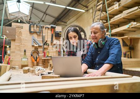 Ein erfahrener Handwerker führt einen jungen Lehrling in einem Holzhof. Sie arbeiten über einen Laptop zusammen, der von Holzbearbeitungswerkzeugen umgeben ist, und betonen dabei die Mentorität Stockfoto