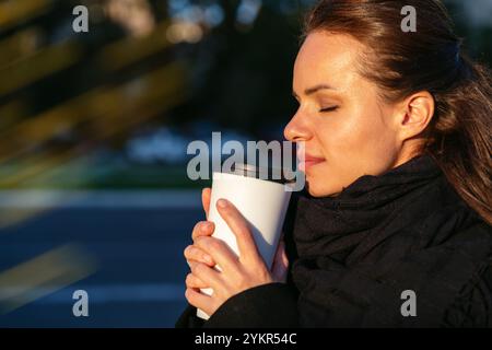 Seitenansicht Porträt einer städtischen Frau mit geschlossenen Augen, die einen Kaffeetasse hält und das Aroma genießt, während sie draußen auf einer Stadtstraße steht. Stockfoto