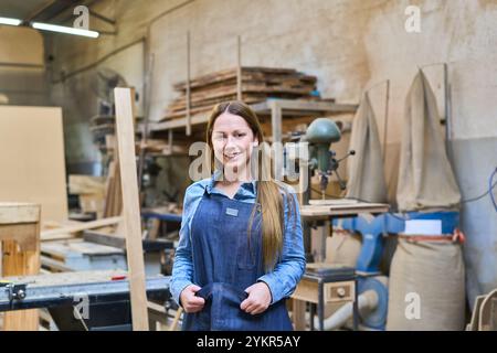 Eine junge Frau posiert selbstbewusst in einem Holzhof, trägt eine Denimschürze. Umgeben von Holz und Werkzeugen verkörpert sie Handwerkskunst und Hingabe in einem W Stockfoto