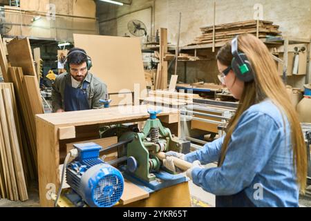 Ein junger Mann und eine junge Frau arbeiten in einer Holzwerkstatt zusammen und konzentrieren sich auf Maschinenbedienung und Teamarbeit. Sie tragen Schutzausrüstung, die auf Sicherheit, Stockfoto