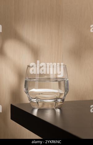 Ein klares Glas Trinkwasser steht auf einem Holztisch, reflektiert das Licht und wirft einen subtilen Schatten, was die Einfachheit und Reinheit des sc unterstreicht Stockfoto