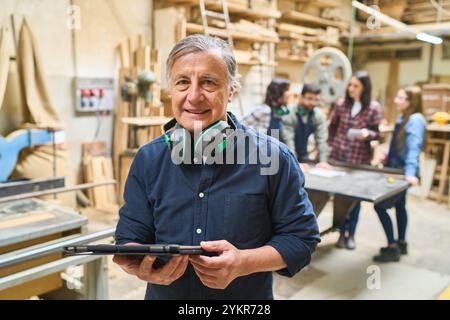 Ein leitender Mitarbeiter hält ein Tablet in einem belebten Holzhof, während jüngere Teammitglieder im Hintergrund zusammenarbeiten und dabei Teamarbeit und Ausbildung hervorheben Stockfoto