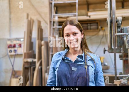 Eine junge Frau in Denim-Hemd und Schürze lächelt selbstbewusst in einer Holzwerkstatt, umgeben von Holzplanken und Maschinen, die Fachkräfte repräsentieren Stockfoto