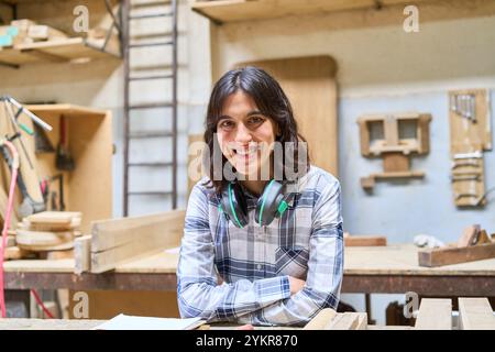 Eine junge Frau lächelt selbstbewusst in einer Holzwerkstatt und zeigt Handwerkskunst und Kreativität. Mit Kopfhörern verkörpert sie einen modernen Ansatz Stockfoto