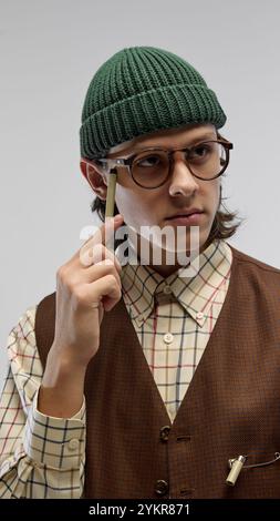 Nahaufnahme eines jungen Mannes in trendiger Brille und grüner Mütze, mit kariertem Hemd im Retro-Stil und brauner Weste, posiert vor dem Lichtstudio Stockfoto