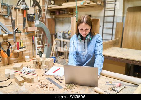 Eine junge Zimmerfrau arbeitet an einem Laptop in einem geschäftigen Holzladen. Umgeben von Werkzeugen und Holzstücken verbindet sie Handwerkskunst mit Technologie, EM Stockfoto