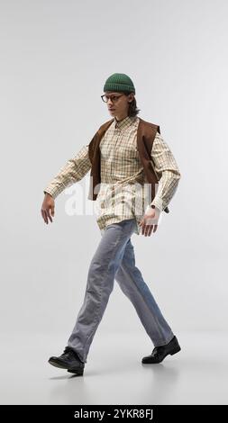 Hübscher junger Mann in grüner Mütze, Brille, kariertem Hemd im Retro-Stil und brauner Weste, posiert in Bewegung vor hellem Studiohintergrund Stockfoto