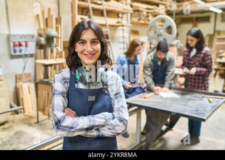 Eine junge Frau steht selbstbewusst in einem belebten Holzhof und zeigt Teamarbeit und Lehre. Im Hintergrund arbeitet eine gemischte Altersgruppe o zusammen Stockfoto