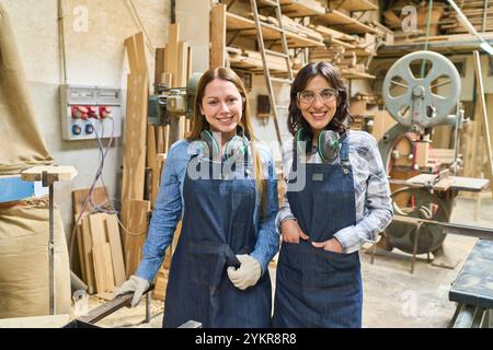Zwei Frauen in einer Holzwerkstatt, die Schutzkleidung und Denim-Schürzen tragen und Teamarbeit und die Entwicklung von Fähigkeiten in einer Holzbearbeitungsumgebung zeigen. P Stockfoto