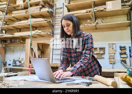 Eine Frau in einem karierten Hemd arbeitet an einem Laptop in einer geschäftigen Holzwerkstatt, umgeben von Holzwerkzeugen und -Materialien, verkörpert Hingabe und Stockfoto