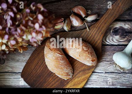 Rustikale Brötchen auf einem Holzbrett mit Knoblauchzehen und getrockneten Blumen im Hintergrund. Gemütliche Komposition im Bauernhausstil. Stockfoto