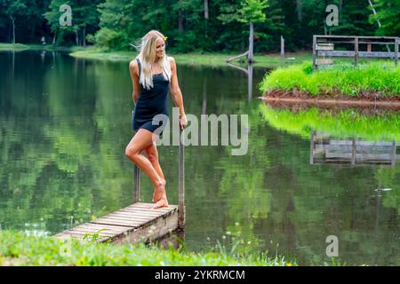 Eine schöne blonde Frau in einem schwarzen Kleid posiert anmutig an einem ruhigen Teich, dessen ruhiges Wasser das üppige Grün um sie herum reflektiert. Die friedliche Lage Stockfoto