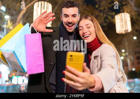 Glückliches Paar, das nachts ein Selfie mit Einkaufstaschen auf der festlichen Stadtstraße macht Stockfoto