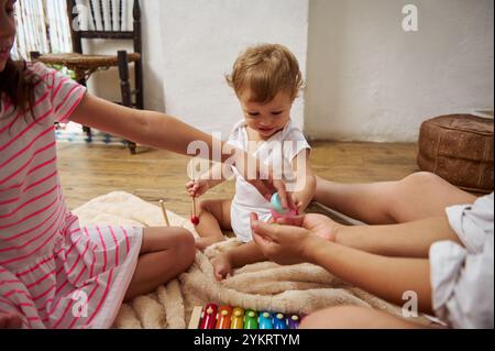 Ein kleines Kind spielt fröhlich mit Geschwistern auf einem Plüschteppich, umgeben von Holzspielzeug und gemütlicher Wohnatmosphäre, was eine herzerwärmende Familienatmosphäre schafft Stockfoto