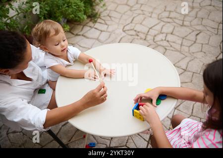 Eine Familie genießt eine schöne Zeit im Freien, während eine Mutter mit ihrem Baby interagiert, während ein anderes Kind mit bunten Blöcken spielt. Die Umgebung ist lässig, gemütlich Stockfoto