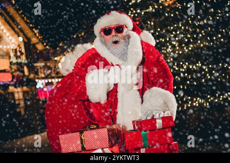 Foto von funky aufgeregt erstaunt santa Carry präsentiert Tasche Stapel geschenkbox Girlande Dekor Neujahr Baum im Freien Stockfoto