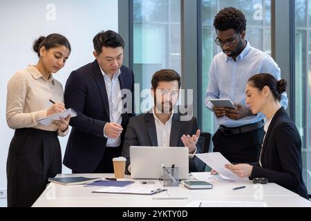 Professionelles Business-Team, das sich an einer gemeinsamen Besprechung rund um Laptop beteiligt und Strategien diskutiert. Die Gruppe umfasst verschiedene Personen mit Dokumenten, Notizblöcken und Tablets, wodurch die Teamarbeit verbessert wird Stockfoto