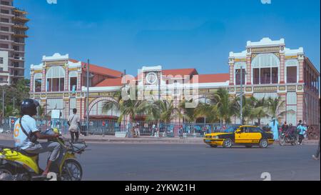 Der Bahnhof Dakar, eine kunstvolle Fassade und ein rotes Dach, umgeben von Palmen. Vor dem Gebäude befindet sich eine belebte Straße mit Fahrzeugen, Stockfoto