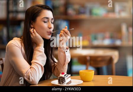 Glückliche arabische Dame, die Dessert und Kaffee mit geschlossenen Augen genießt, im Café sitzt und bei der Arbeit Pause macht Stockfoto