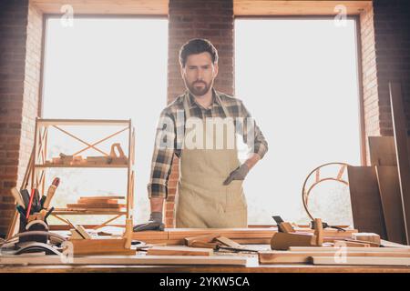 Foto eines starken, selbstbewussten bärtigen Mannes mit spezieller Schürze und Handschuhen, der in der Nähe seiner Handwerker oder hölzernen Flugzeugmodelle posiert Stockfoto