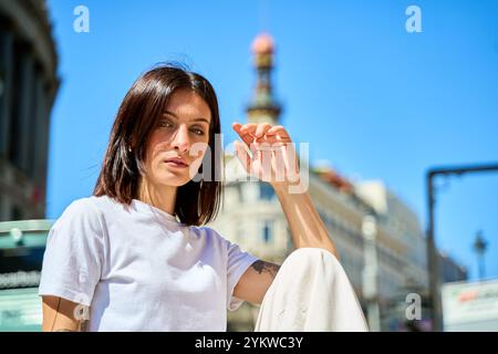 Eine junge Frau, die auf einem Steinvorsprung in einer Stadtstraße sitzt und ihre Augen mit einer Hand vor der Sonne abschirmt, mit einer grünen Handtasche neben ihr. Stockfoto