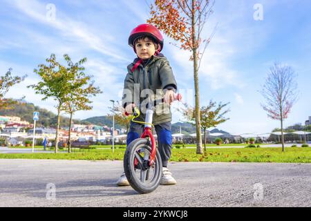 Ein kleiner Junge fährt mit Helm auf dem Fahrrad. Die Szene spielt in einem Park mit Bäumen im Hintergrund. Der Junge scheint sich zu amüsieren, wie er Stockfoto