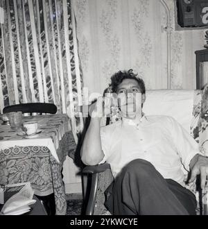 1960er Jahre, historisch, häusliches Leben, ein Mann Anfang dreißig, der nach der Arbeit in einem hölzernen Sessel drinnen sitzt, neben einem mit Stoff bedeckten Tisch, eine Pfeife raucht, rechte Hand auf dem Pfeifenrohr, England, Großbritannien. Stromzähler der Ära an der Wand dahinter. Stockfoto