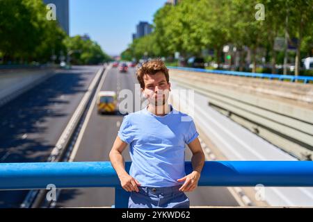 Ein Mann in einem blauen Hemd steht auf einer Brücke mit Blick auf eine geschäftige Straße. Er sieht gelangweilt aus und interessiert sich nicht für die Szene Stockfoto