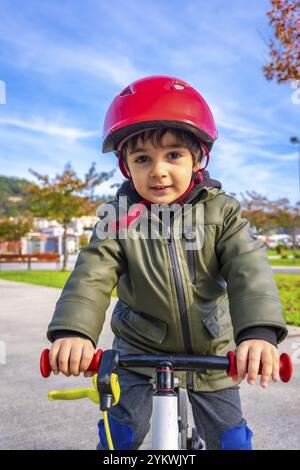 Ein kleiner Junge mit grüner Jacke und rotem Helm fährt Fahrrad. Der Junge lächelt und er genießt sich Stockfoto