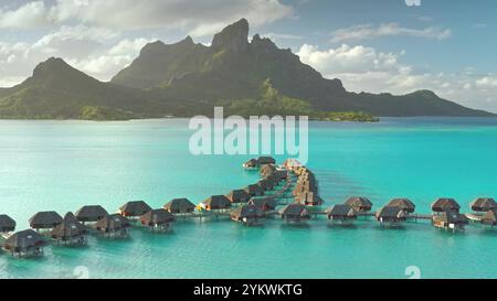 Die Bungalows aus der Vogelperspektive über dem Wasser bilden sich in Richtung Mount Otemanu auf der exotischen Insel Bora Bora, mit ihrer türkisfarbenen Lagune und üppiger Vegetation. Luxuriöses Reiseurlaubsresort im Korallenriffmeer Stockfoto