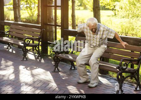 Senior-Mann hält sich den Rücken. Alter Mann in der Nähe der Parkbank. Starke Schmerzen in der Wirbelsäule. Medikamentenbedarf und Therapie Stockfoto