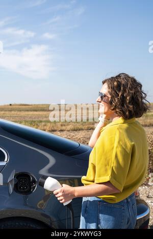 Eine Frau mit gewölbten Haaren, die ein gelbes Hemd und eine Sonnenbrille trägt, steht neben dem Aufladen des Elektroautos. Windkraftanlage im Hintergrund. Stockfoto