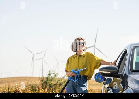 Die Frau hält eine Wasserstoff-Tankdüse. Betanken des Autos mit Wasserstoffkraftstoff. Windräder im Hintergrund. Stockfoto