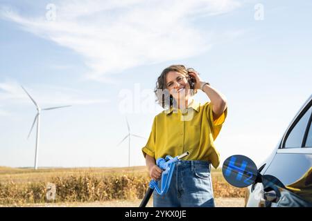 Die Frau hält eine Wasserstoff-Tankdüse. Betanken des Autos mit Wasserstoffkraftstoff. Windräder im Hintergrund. Stockfoto