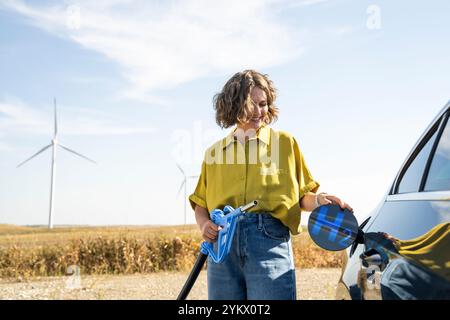 Die Frau hält eine Wasserstoff-Tankdüse. Betanken des Autos mit Wasserstoffkraftstoff. Windräder im Hintergrund. Stockfoto