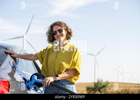 Die Frau hält eine Wasserstoff-Tankdüse. Betanken des Autos mit Wasserstoffkraftstoff. Windräder im Hintergrund. Stockfoto