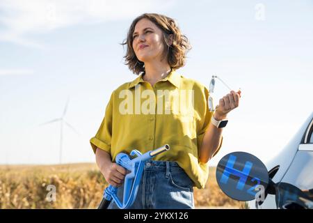 Die Frau hält eine Wasserstoff-Tankdüse. Betanken des Autos mit Wasserstoffkraftstoff. Windräder im Hintergrund. Stockfoto