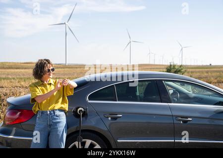 Eine Frau mit gewölbten Haaren, die ein gelbes Hemd und eine Sonnenbrille trägt, steht neben dem Aufladen des Elektroautos. Windräder im Hintergrund. Stockfoto