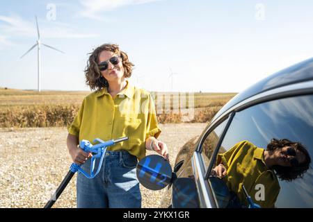 Die Frau hält eine Wasserstoff-Tankdüse. Betanken des Autos mit Wasserstoffkraftstoff. Windräder im Hintergrund. Stockfoto