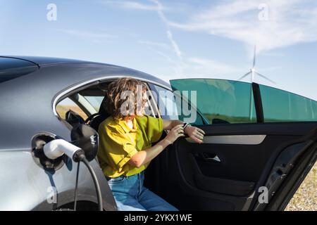 Eine Frau mit gebogenen Haaren, die ein gelbes Hemd trägt, sitzt in einem Elektroauto. Windräder im Hintergrund. Stockfoto