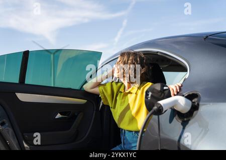 Eine Frau mit gebogenen Haaren, die ein gelbes Hemd trägt, sitzt in einem Elektroauto. Windräder im Hintergrund. Stockfoto